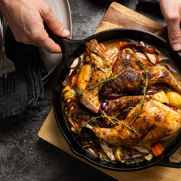 Hands holding a cast iron skillet of rabbit stew with root vegetables and fresh herbs on a wood cutting board.